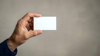 Male hand holding blank white business card against neutral gray background for corporate branding and professional networking purposes.