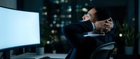Stressed businessman working late at night in modern office, holding head in hands showing burnout and exhaustion from overtime work pressure.