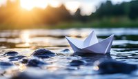 White paper boat floating on rippling water surface at sunset with golden bokeh lights reflecting on waves creating dreamy atmosphere.