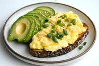 Creamy scrambled eggs on dark bread topped with green onions, served alongside fresh sliced avocado on gray ceramic plate against white background.