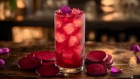Refreshing red berry cocktail with ice cubes in tall glass surrounded by purple flower petals on rustic wooden table with warm bokeh background lighting.