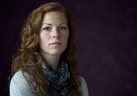 Young woman with long wavy auburn hair wearing casual white sweater and teal scarf gazes thoughtfully against dark purple background in dramatic studio lighting.