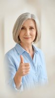 Mature woman with gray hair wearing light blue shirt giving thumbs up gesture, expressing approval and positivity in studio setting.