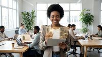 Young African American woman with natural curly hair wearing a beige cardigan, holding a tablet in a bright modern office with colleagues working at desks and green plants.