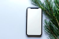 Modern smartphone with blank white screen mockup surrounded by fresh green pine branches on light background for holiday app presentation and digital marketing.