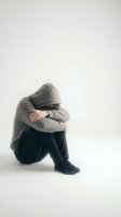 Depressed young man in hoodie sitting alone with head down expressing sadness, mental health struggles and emotional pain on white background.