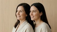 Two Caucasian women, senior mother and adult daughter, smiling together against a light wooden background, showing family bond and generational connection.