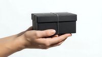 Black gift box with silver ribbon held in cupped hands against white background, isolated for design use in greeting cards and promotional materials.