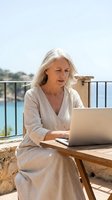 Mature woman with white hair, Caucasian ethnicity, working on laptop at outdoor terrace overlooking turquoise water. Remote work, freelance, digital nomad lifestyle concept for business and technology use.