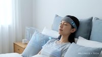 Young Asian woman relaxing in bed wearing blue sleep tracking headband device for wellness monitoring and health technology applications.