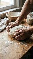 Hands kneading fresh bread dough on wooden cutting board in rustic kitchen with flour and baking ingredients for homemade artisan bakery preparation.
