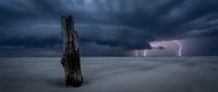 Weathered wooden post stands alone on vast sandy beach under dramatic stormy sky with lightning striking distant horizon during tempest.