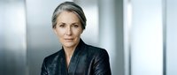 Mature woman with gray hair wearing dark blazer in modern office setting, professional portrait for corporate business use.