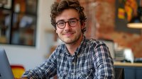 Young Caucasian male professional with glasses and curly hair wearing plaid shirt smiling while working on laptop in modern cozy cafe. Perfect for business, freelance, and lifestyle content.
