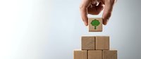 Hand placing wooden block with green tree icon on top of stacked cubes representing sustainable business growth and environmental responsibility.
