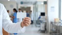 Doctor in white coat holding blank business card in hospital clinic setting with blurred medical staff and equipment in background for healthcare branding and professional identification.