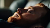 Young caucasian man with red beard sleeping peacefully in dark room with dramatic side lighting creating moody atmosphere and deep shadows on face.