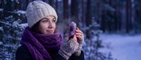Young Caucasian woman in knit hat and purple scarf warming hands in snowy winter forest, breath visible in cold air.