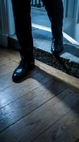 Man in formal black suit and polished leather shoes standing at doorway threshold, stepping between indoor wooden flooring and outdoor carpeted entrance. Business professional concept.