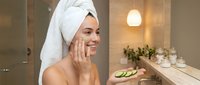 Young woman applying cucumber slices on face in modern bathroom with towel on head for natural skincare routine and spa treatment at home.