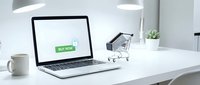 Laptop with buy now button on screen beside miniature shopping cart on white desk, representing online shopping and e-commerce concept.
