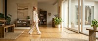 Woman in beige linen clothing walking through bright, minimalist home interior with wooden floors, natural light, and potted plants for lifestyle and wellness content.
