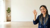Young woman with dark hair and South Asian ethnicity wearing navy blue traditional embroidered clothing, waving hand in greeting gesture at wooden table with potted plant in bright interior space.