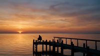 Silhouette of person sitting on wooden pier at sunset over calm water, golden hour reflection on ocean surface.