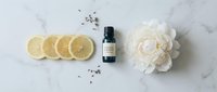 Overhead view of essential oil bottle surrounded by fresh lemon slices, dried lavender buds, and white peony flower on marble surface for wellness and aromatherapy design.