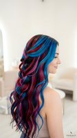 Young woman with long, wavy hair featuring vibrant blue, pink, and burgundy color blocking and braided sections, shown in profile against a neutral background.