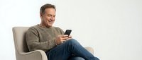 Middle-aged Caucasian man in casual attire sitting in armchair, holding smartphone, smiling while using mobile device for communication, shopping, or browsing online content.