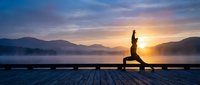 Woman practicing yoga warrior pose on wooden dock at sunrise with mountain lake backdrop and colorful sky for wellness meditation and fitness concepts.