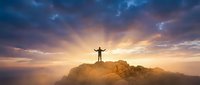 Man with arms raised standing on mountain peak at sunset, celebrating success and achievement with dramatic sky and clouds above.