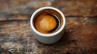 Fresh espresso with golden crema in white ceramic cup on rustic wooden table background, shallow depth of field close-up shot highlighting rich coffee texture.