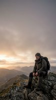 Male hiker in dark winter gear sitting on mountain summit at sunset, overlooking vast valley landscape with distant peaks and dramatic cloudy sky. Adventure and outdoor recreation.