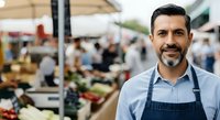 Male vendor with dark hair and beard wearing blue apron at outdoor farmers market stall, suitable for agriculture, small business, and retail concepts.