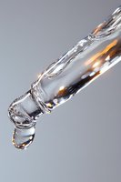 Crystalline water drop forming on transparent icicle with light refraction and golden highlights against soft gray background, macro photography.