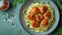 Glazed chicken meatballs served on fettuccine pasta with fresh herbs and spices, garnished with basil leaves on gray ceramic plate against turquoise background.