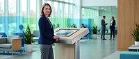 Professional businesswoman using modern office printer in contemporary corporate workspace with glass walls and colleagues in background.