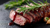 Juicy medium rare beef steak sliced on dark stone background with fresh rosemary sprigs and red wine sauce, showing perfect grill marks and pink center.