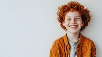 Cheerful redhead Caucasian boy with curly hair smiling broadly, wearing an orange shirt, isolated on a white background.