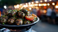 Grilled meatballs garnished with fresh herbs and cherry tomatoes on black plate, evening outdoor dining with festive bokeh lights in background.