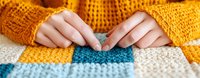 Female hands in orange knitted sweater working on colorful striped crochet project with blue, white and yellow yarn pattern, close-up view of crafting process.