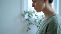 Young Caucasian woman in profile wearing green shirt looking down pensively in bright modern interior with natural light and plants in background.