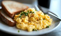 Creamy scrambled eggs garnished with fresh chives and black pepper served alongside toasted bread on white plate, shallow depth of field breakfast photography.