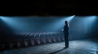 Silhouette of woman standing alone in empty theater auditorium with dramatic stage lighting and rows of seats creating atmospheric performance venue.