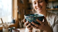 Female potter with paint-stained hands holding handmade ceramic bowl in studio workshop. Artisan craftswoman displaying glazed pottery creation.