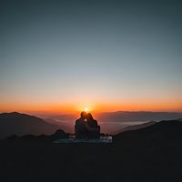 Romantic couple silhouette embracing at mountain summit during golden sunset with layered hills and warm orange sky creating intimate moment.
