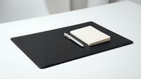Minimalist workspace setup with black desk pad, cream notebook and silver pen on white table surface for professional office environments.