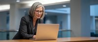 Middle-aged woman with gray hair and glasses wearing black blazer working on laptop at office desk with modern interior background.